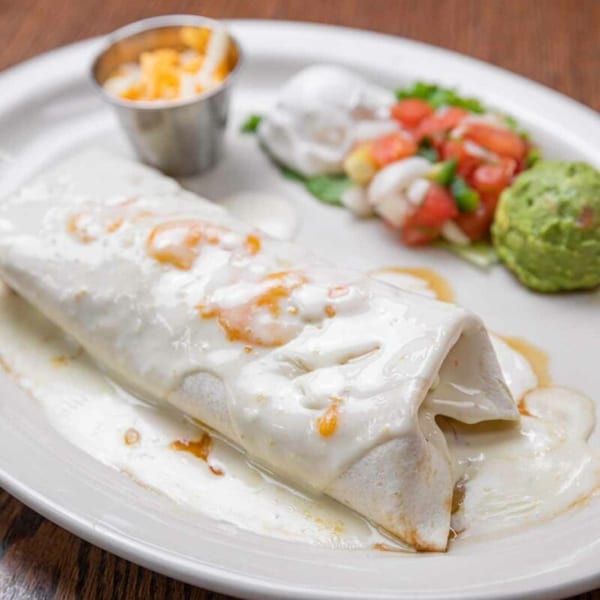 A Pecina's Mexican Cafe burrito covered in white sauce, with sides of guacamole, pico de gallo, and cheese.