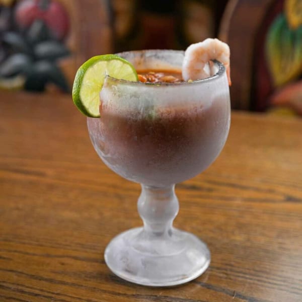 A frosty Pecina's Mexican Cafe michelada with a lime wedge and shrimp on a wooden table.