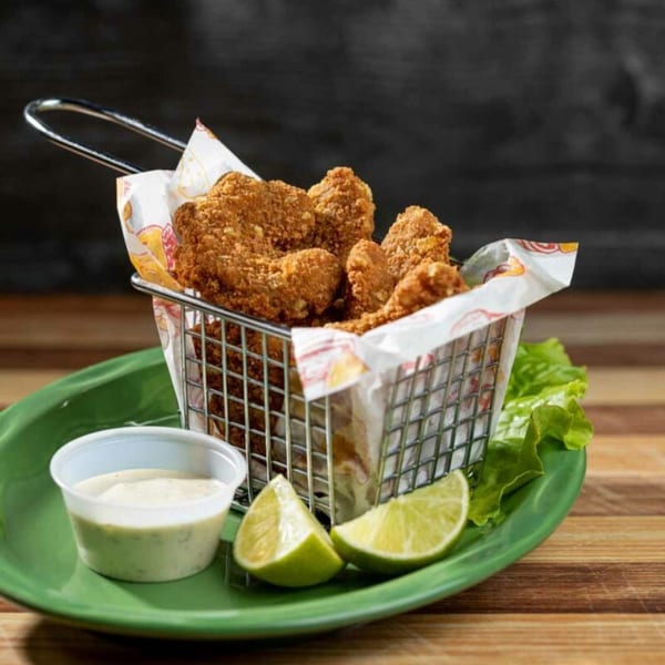 Crispy fried chicken pieces in a metal basket with lime wedges and dipping sauce on a green plate.