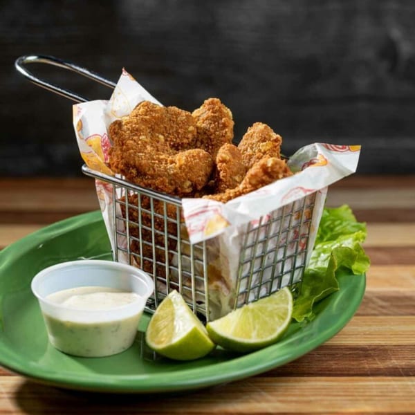 Crispy fried chicken bites in a metal basket with lime wedges and dipping sauce on a green plate.