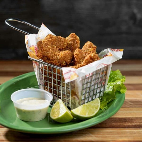 Crispy fried chicken bites in a metal basket with lime wedges and a side of creamy sauce.