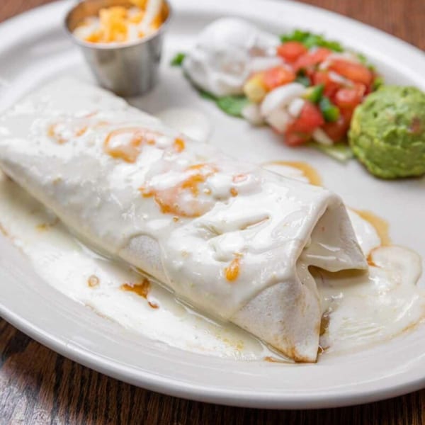 A Pecina's Mexican Cafe burrito covered in white sauce, with sides of guacamole and pico de gallo.